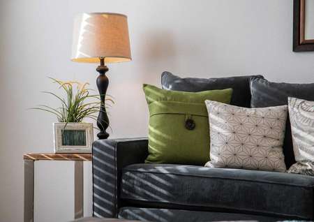 Model home with dark blue sofa that has green and white pillows, framed picture hanging on the wall, and lamp with beige shade in the corner