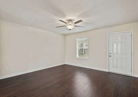 The living room of the Cumberland floor plan is equipped with a ceiling fan and window with 2 inch faux blinds, brown vinyl wood-like flooring, white baseboards and a white door