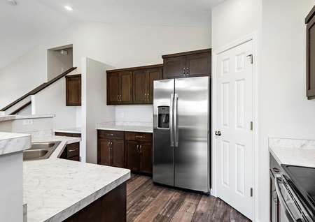 Closeup photo of a kitchen with white counters, brown cabinets, stainless appliances and view of stairs.