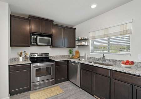 Photo of a kitchen with dark brown cabinets, stainless steel electric range and built-in microwave, speckled gray granite counters and a window over the single-basin sink.