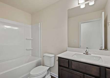 The second full bath from the Cumberland has a tub and shower and white cultured marble countertop with a full mirror and lighting above.  There is a toilet between the sink and the bathtub 