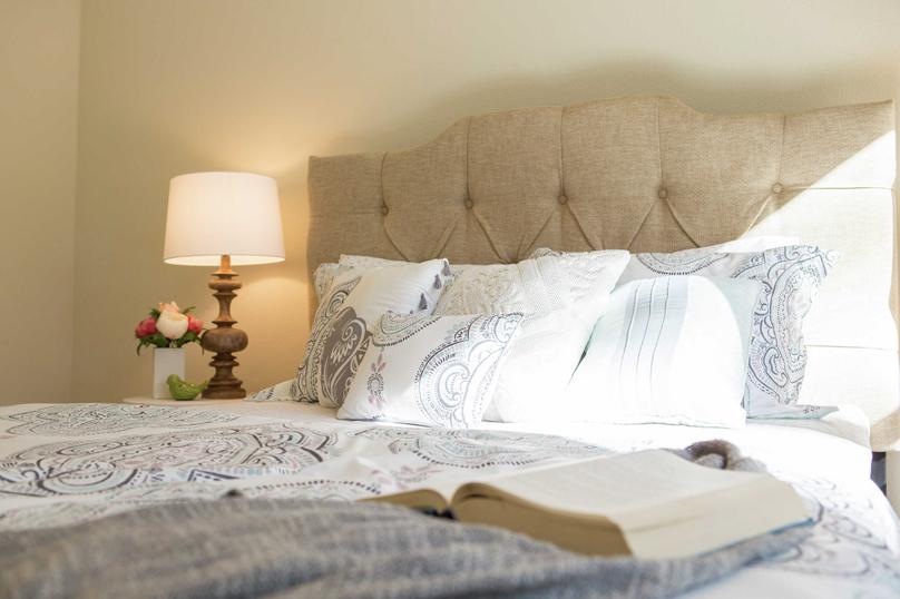 This bedroom is staged with a sandy color button tufted headboard, open book on top of a bluish comforter with white pillows on the bed, and a lit lamp with beige shade