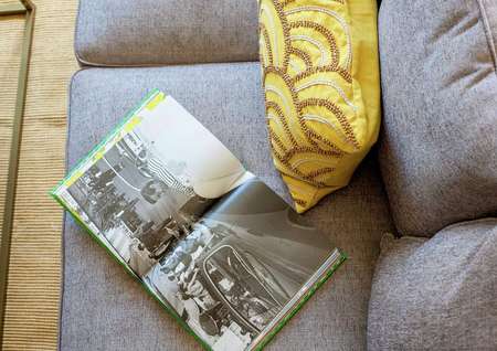 Staged living room with grey sofa, yellow throw pillow with brown pattern, and open book with black and white picture on it