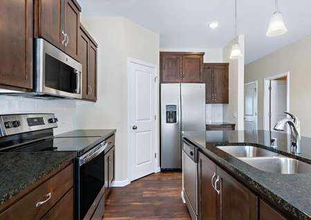 Photo of kitchen with brown cabinets, dark gray granite counters, stainless appliances and brown plank flooring.