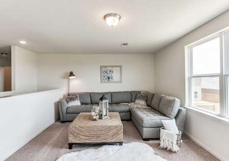 Driftwood staged loft in model with grey sectional sofa, white throw rug, and wooden wicker coffee table with short black legs