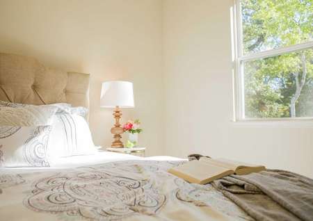 Staged bedroom with bed that has a white comforter, brown blanket, and open book on it plus a bedside table with a white-shade lamp and vase with flowers