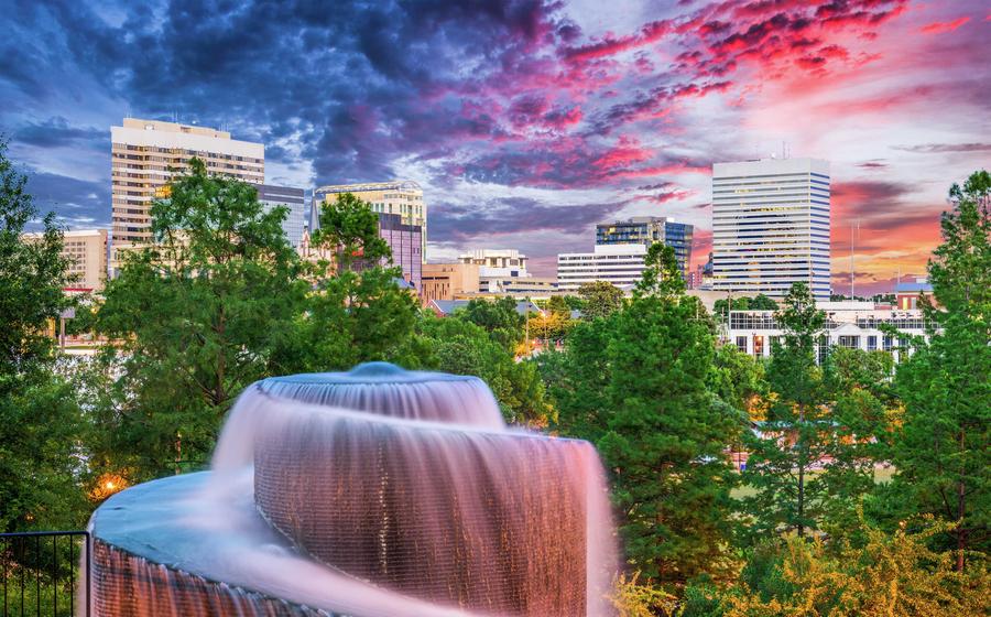 Columbia, South Carolina spiralling fountain surrounded by green pines with downtown tall buildings and multicolor pink, blue, and orange sky