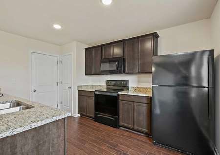 Image of the Cumberland plan’s kitchen with granite countertops, black appliances, dark brown cabinetry, stainless steel sink and wood-style vinyl flooring