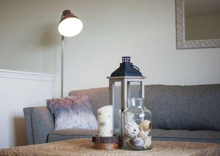 Driftwood staged home living room with gray sofa with throw pillow, standing lamp with light on in the corner, and decorative candle and lantern on a wood table