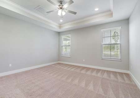 Spacious master bedroom with two windows, a fan and tan walls and carpet.