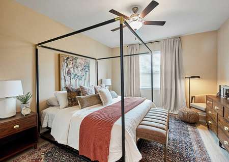 Photo of decorated master bedroom with iron four-post bed with earth tone bedding, a window with drapes, ceiling fan and carpet.
