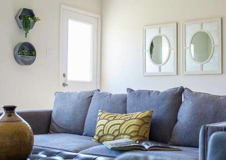 Model home staged with blue sofa with brown pattern throw pillow, mirror mounted on the wall, and decorative plants on the other wall