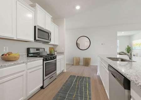 Staged kitchen with white cabinets, plank flooring, stainless range with overhead microwave, granite counters, runner rug, into foyer with circle mirror, stools, view of dining room with artwork and tall plant in corner.