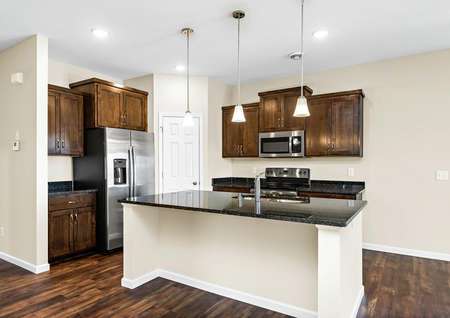Photo of an open kitchen with brown cabinets, dark gray granite counters, stainless steel appliances and brown plank flooring.