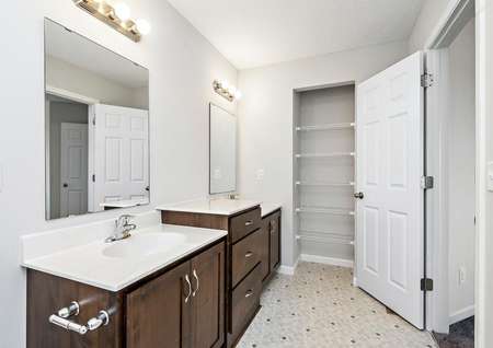 Photo of a spacious bathroom with a split vanity, brown cabinets with silver hardware and view into a linen closet.