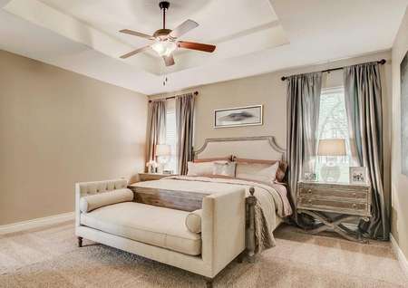 Staged master bedroom with king bed, taupe and silver decor, two windwos with gray silk drapes, upholstered bench, double nightstands and lamps, ceiling fan, coffered ceiling.