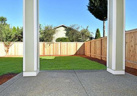 Photo taken from a covered back patio overlooking a back yard with a wooden fence, green grass and a perimeter border of mulch inside the fence.