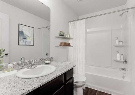 Staged bathroom with single sink vanity, toilet and tub-shower combo with white shower curtain, dark flooring and frameless mirror.