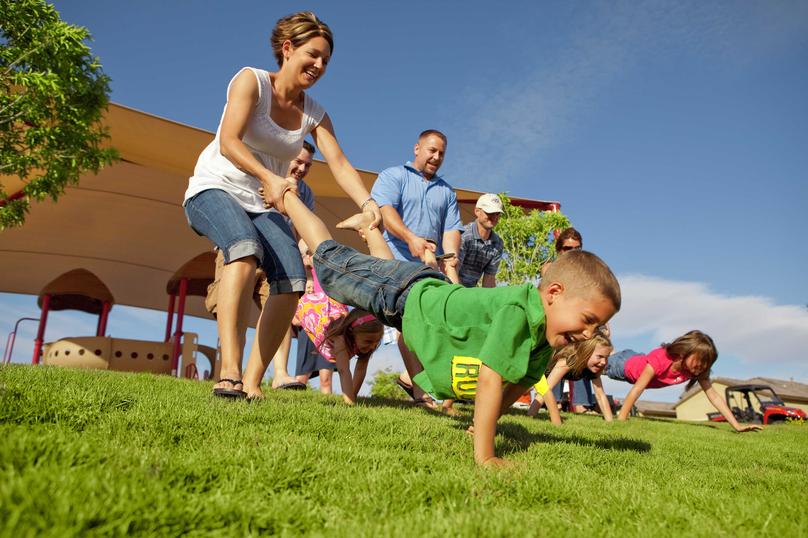 Families playing wheelbarrow races on a grassy lawn.