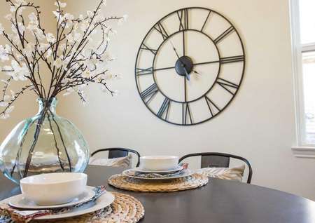 Maple model home dining nook staged with wooden table with placemats, white bowels, and glass ornament with flowers plus large wall clock in the back