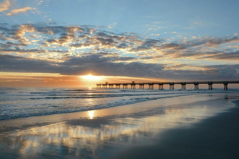 Jacksonville, Florida beach sunrise with pier extended far into the ocean, rolling waves, and sunlight reflecting off the wet sand
