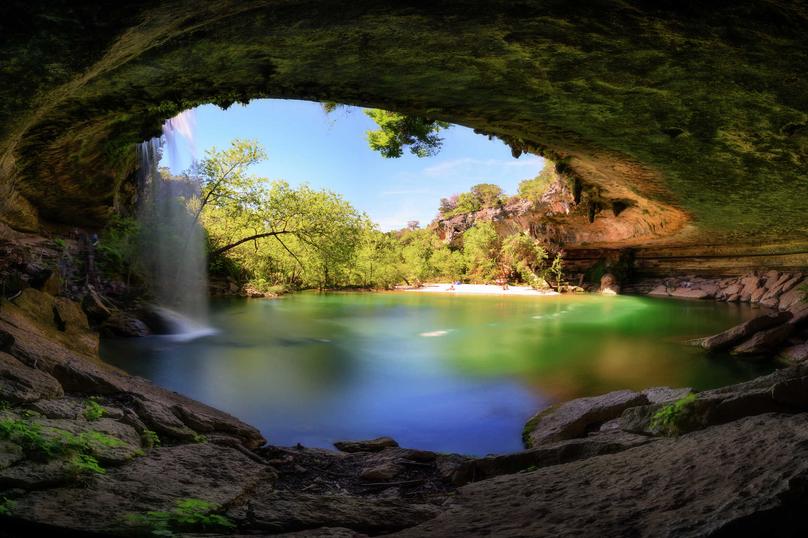 Austin, Texas Hamilton Pool waterfall recreational area with stone cave, water falling over the cliff, and trees off in the distance
