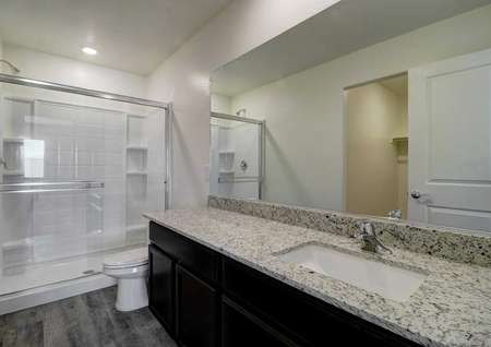 A bathroom in the Cottonwood floor plan granite countertops, dark brown cabinets and a walk-in shower.
