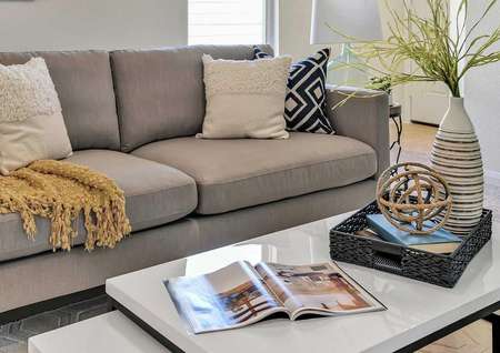 Staged home with gray sofa with white and gray and black pillows, coffee table with open magazine and decoration and wall art hanging on the wall